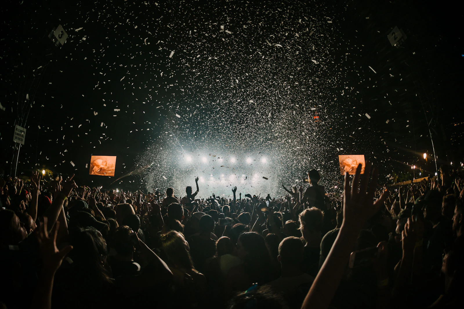 Crowd at an open-air concert facing an outdoor festival stage with lights and confetti